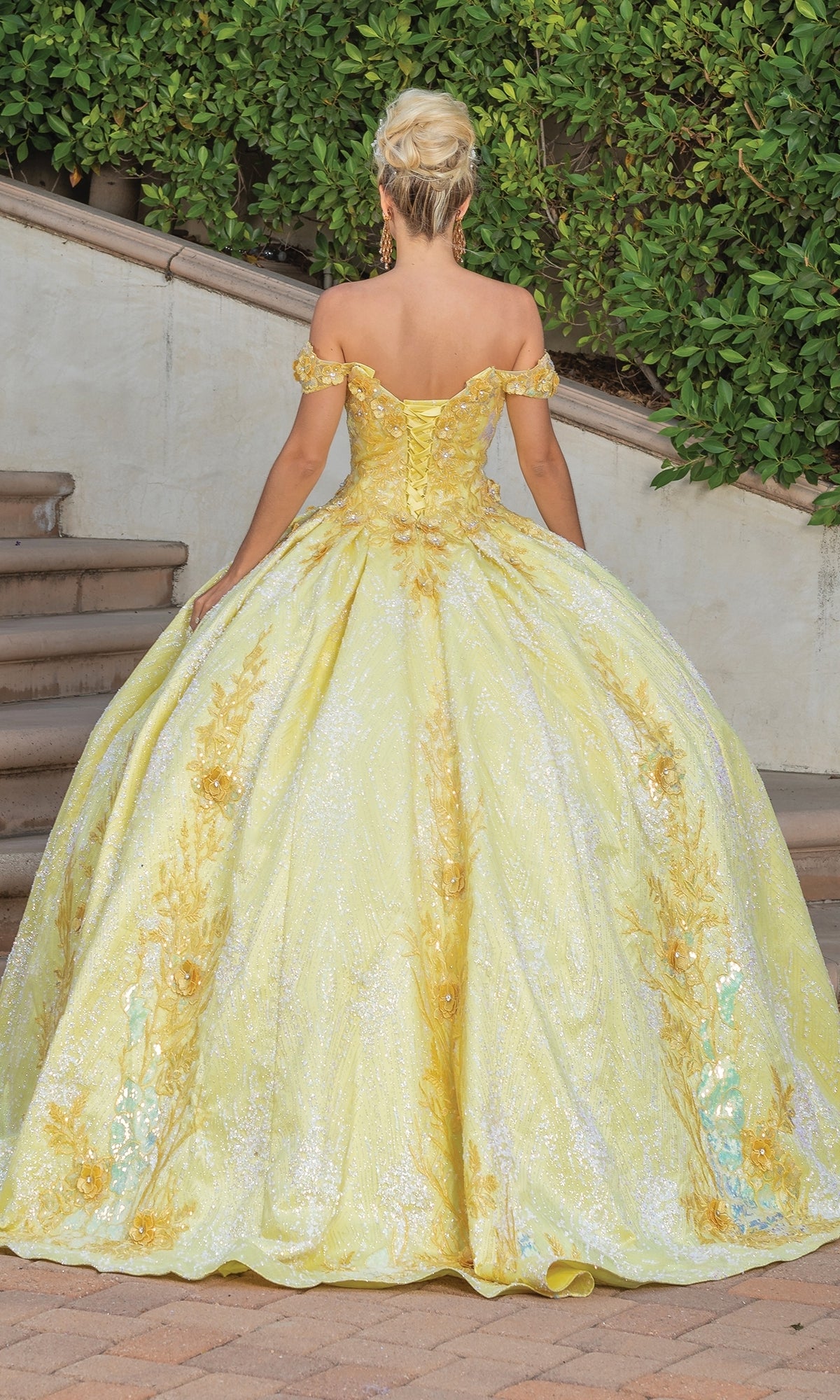 A woman stands outdoors on stone steps, facing away, wearing the Quinceanera Dress 1772 By Dancing Queen—an a-line ball gown with off-the-shoulder sleeves, sparkling yellow fabric, and floral details.